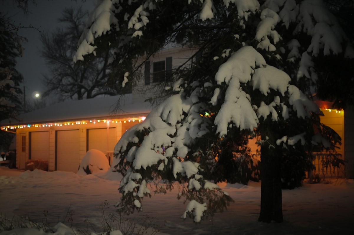 Snow-covered home with a lit tree glowing in the yard at night